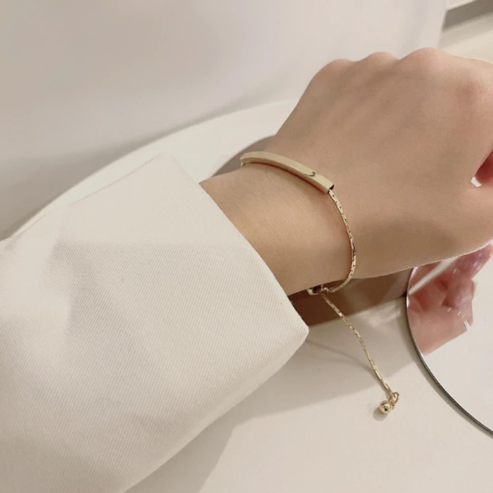 Hand wearing a gold bracelet with a dangling chain, against a white fabric background.