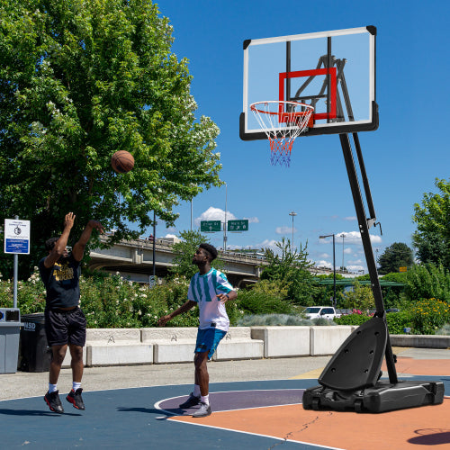 Two people play basketball on a sunny outdoor court with a blue and orange surface near greenery.