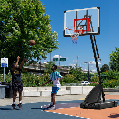 Two people play basketball on a sunny outdoor court with a blue and orange surface near greenery.