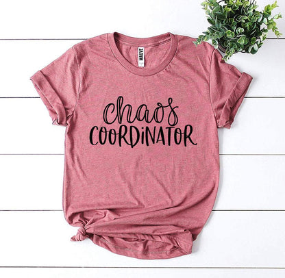 Pink T-shirt with "chaos coordinator" text in black. Plant in top right corner. White background.