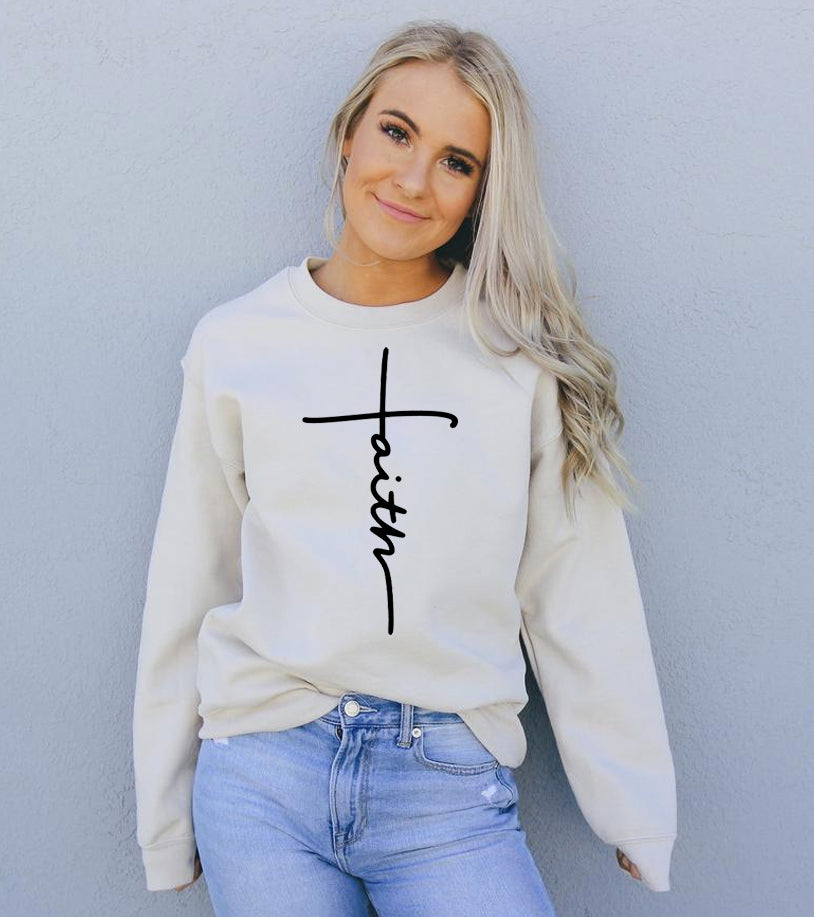 Woman in cream sweater with "faith" in black text, wearing light blue jeans, standing by a gray wall.