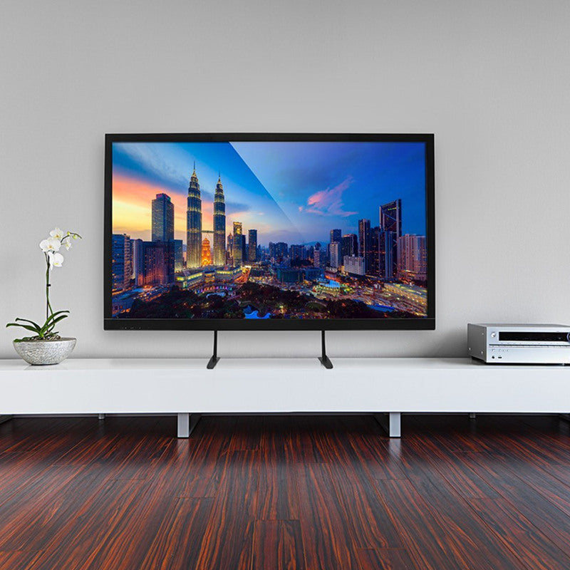 TV on a white stand, displaying a cityscape at sunset. Wooden floor, gray wall, and a white orchid.