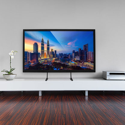 TV on a white stand, displaying a cityscape at sunset. Wooden floor, gray wall, and a white orchid.
