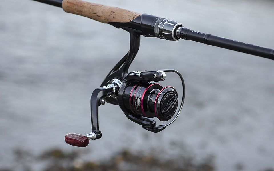 Close-up of a fishing rod with a black reel, red accents, and a cork handle by water.