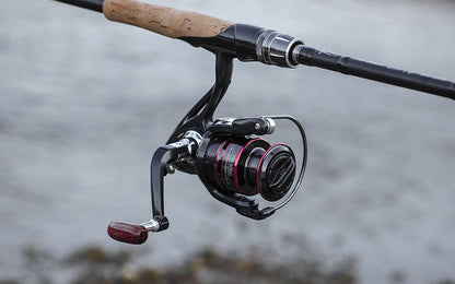 Close-up of a fishing rod with a black reel, red accents, and a cork handle by water.