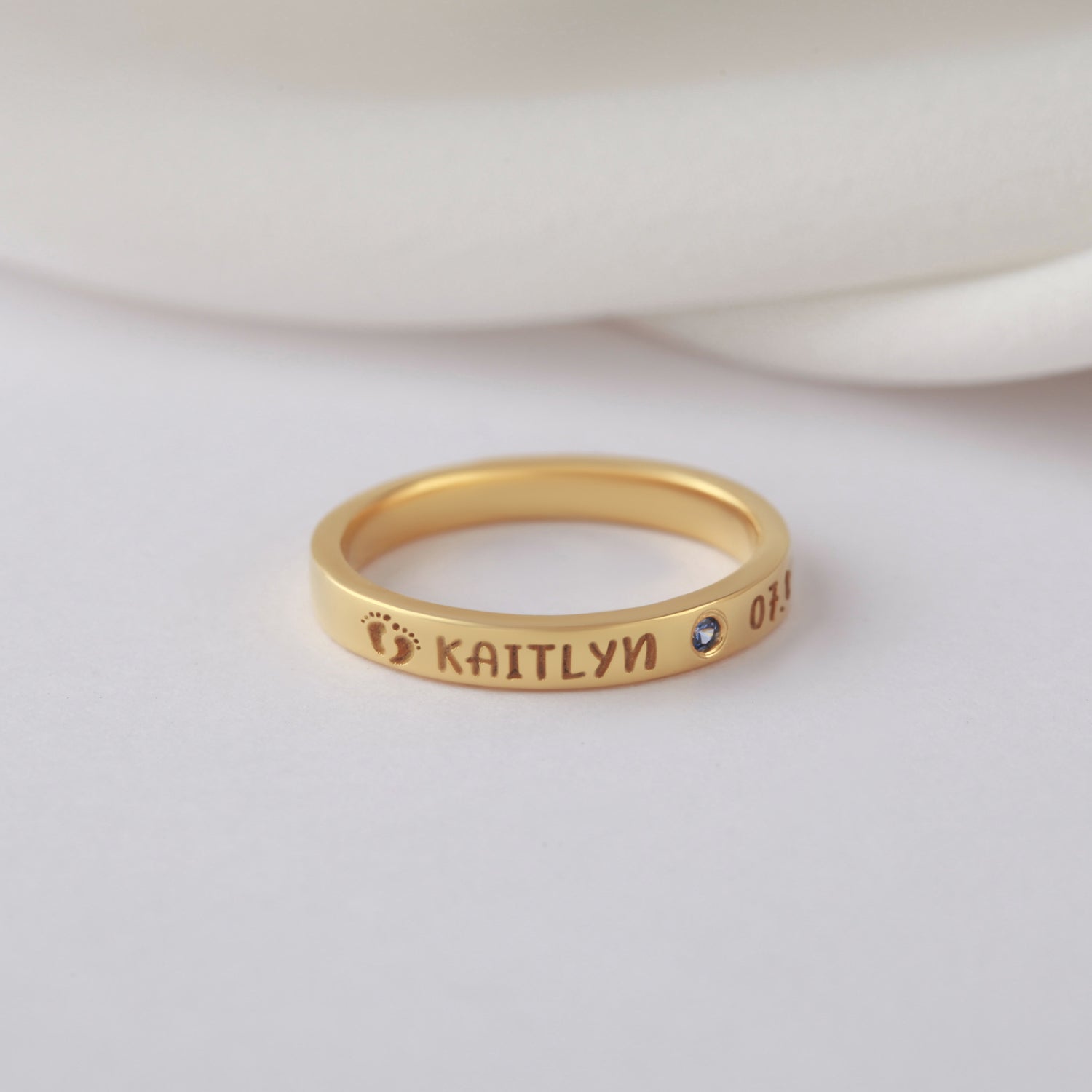 Gold ring with engraved name, blue gemstone, tiny footprint, and numbers on a soft background.