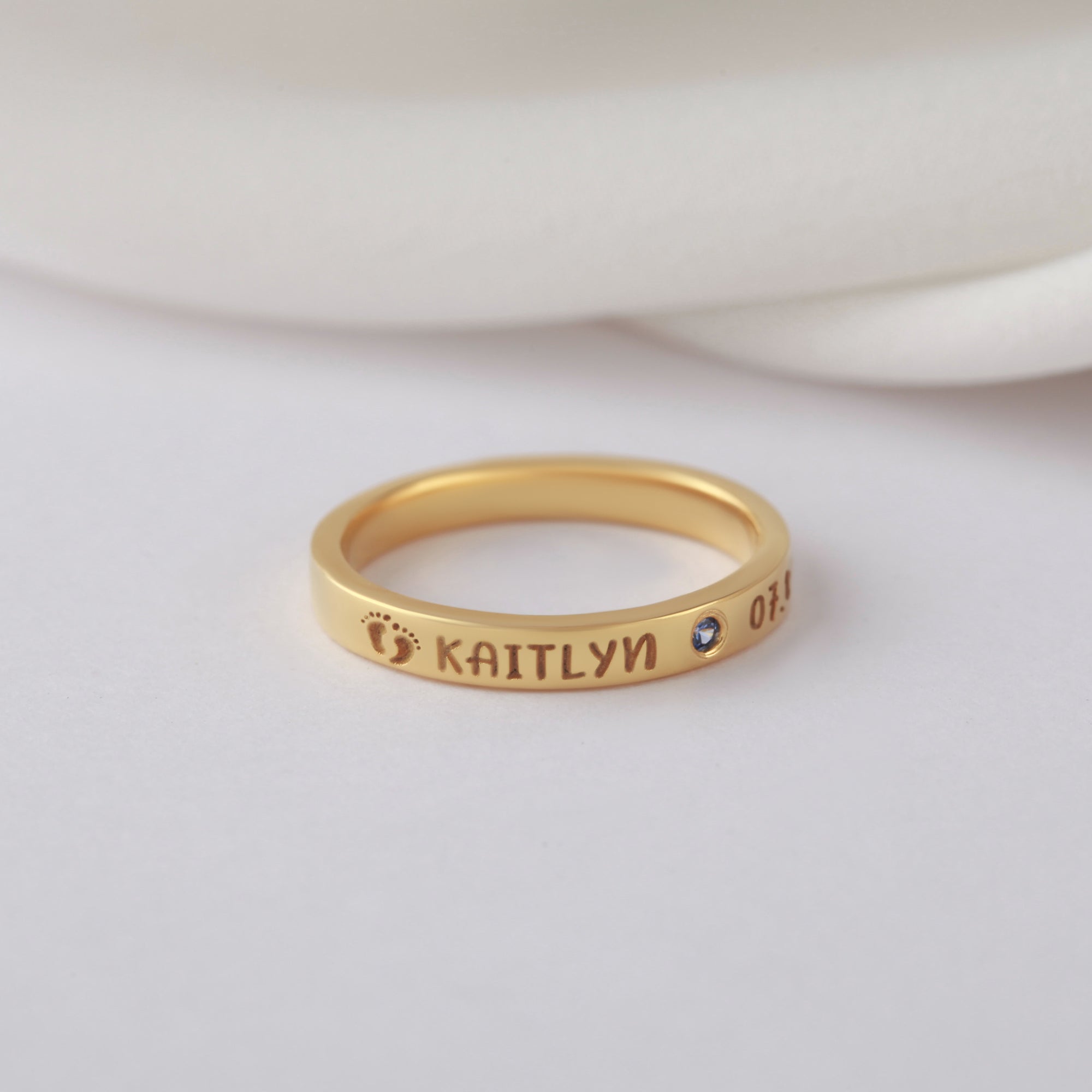 Gold ring with engraved name, blue gemstone, tiny footprint, and numbers on a soft background.