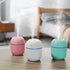 Three colorful humidifiers—pink, white, and teal—emitting mist on a wooden table.