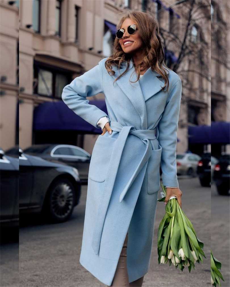 Woman in blue coat holding white tulips, smiling, wearing sunglasses on a city street.