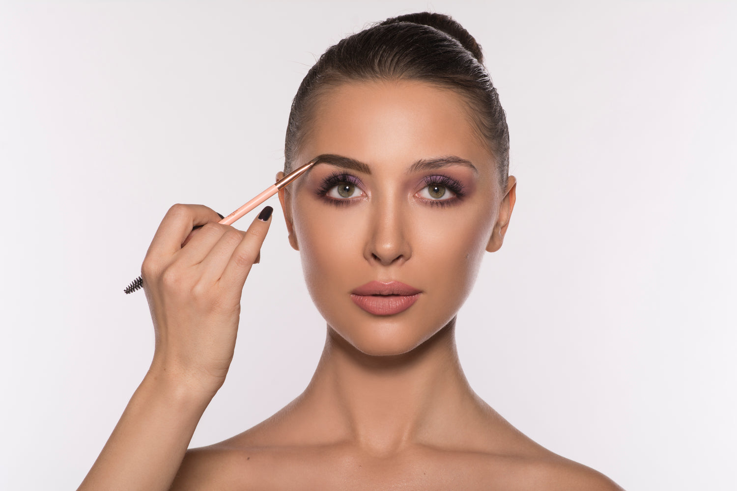 Woman applying makeup with brow pencil, pink eyeshadow, and nude lips on a light background.