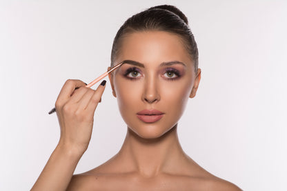 Woman applying makeup with brow pencil, pink eyeshadow, and nude lips on a light background.