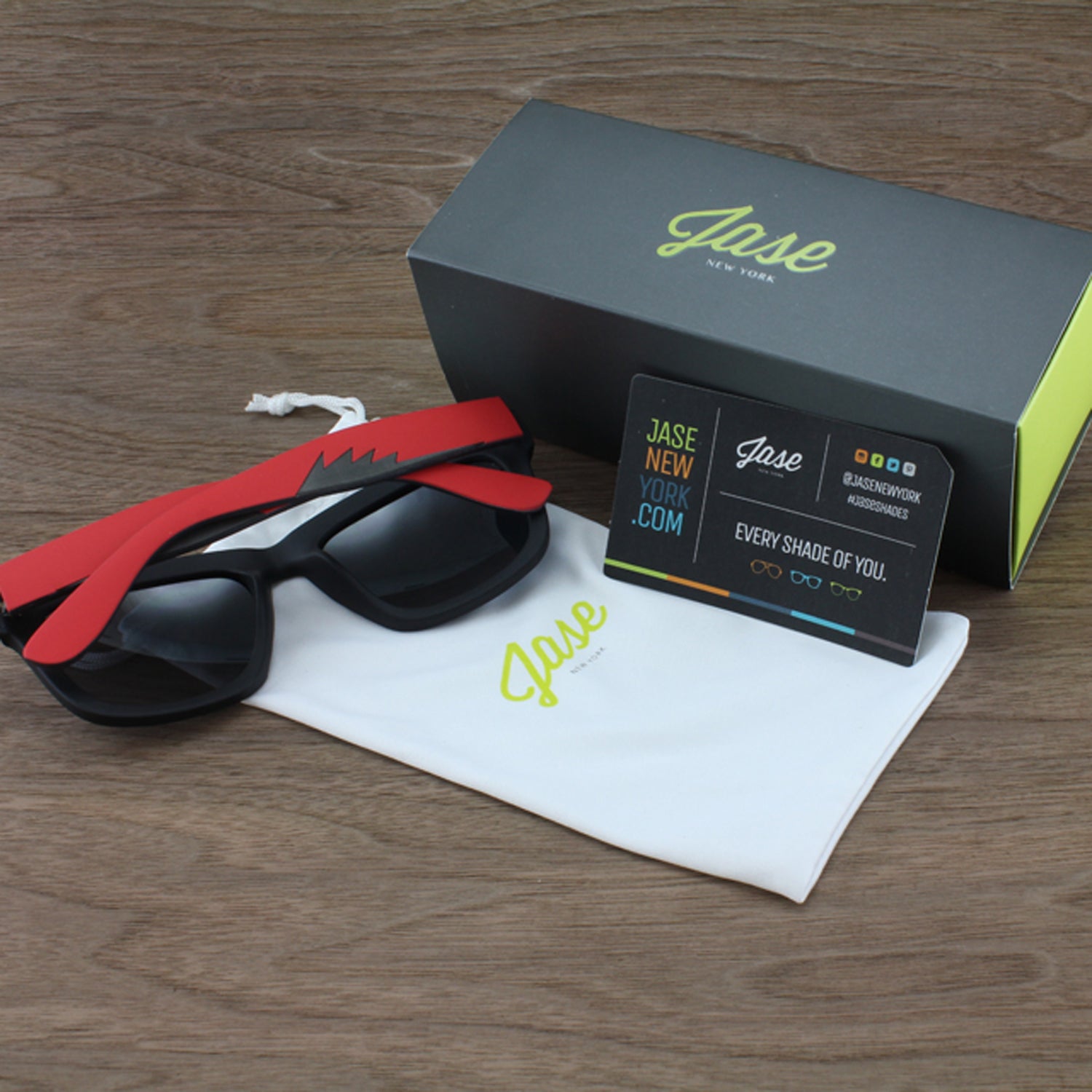 Sunglasses with red and black frames, white pouch, black box, and card on a wooden surface.