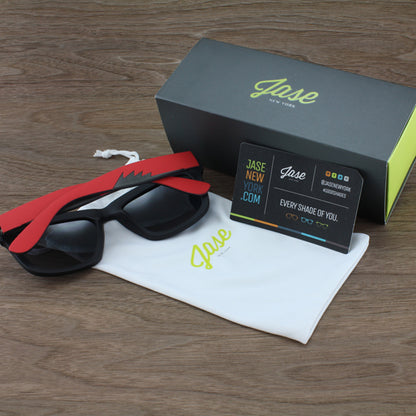 Sunglasses with red and black frames, white pouch, black box, and card on a wooden surface.