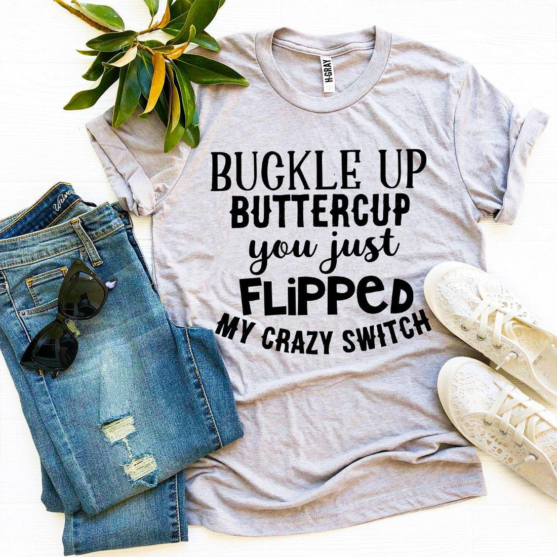 Gray T-shirt with bold black text beside ripped jeans, sunglasses, white shoes, and green leaves.