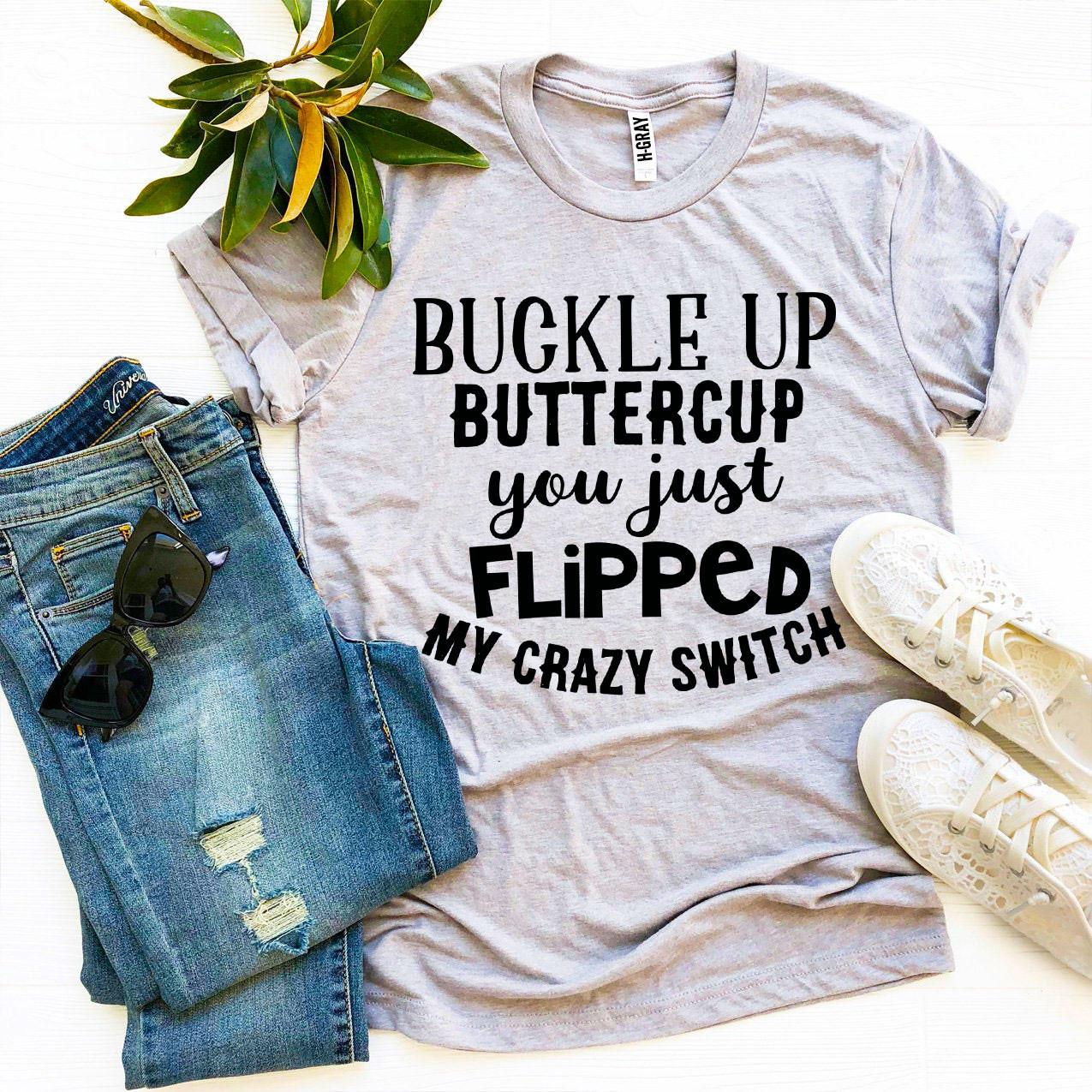 Gray T-shirt with bold black text beside ripped jeans, sunglasses, white shoes, and green leaves.