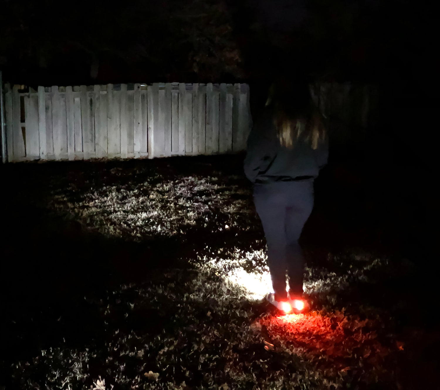 Person in dark clothing walking at night, illuminated by glowing red shoes near a wooden fence.