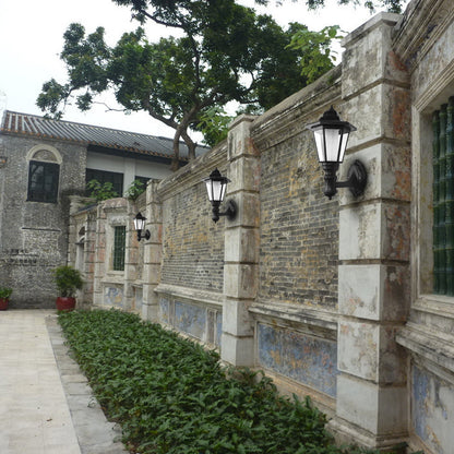 Old stone wall with black lanterns, green foliage, and a building with tiled roof in the background.