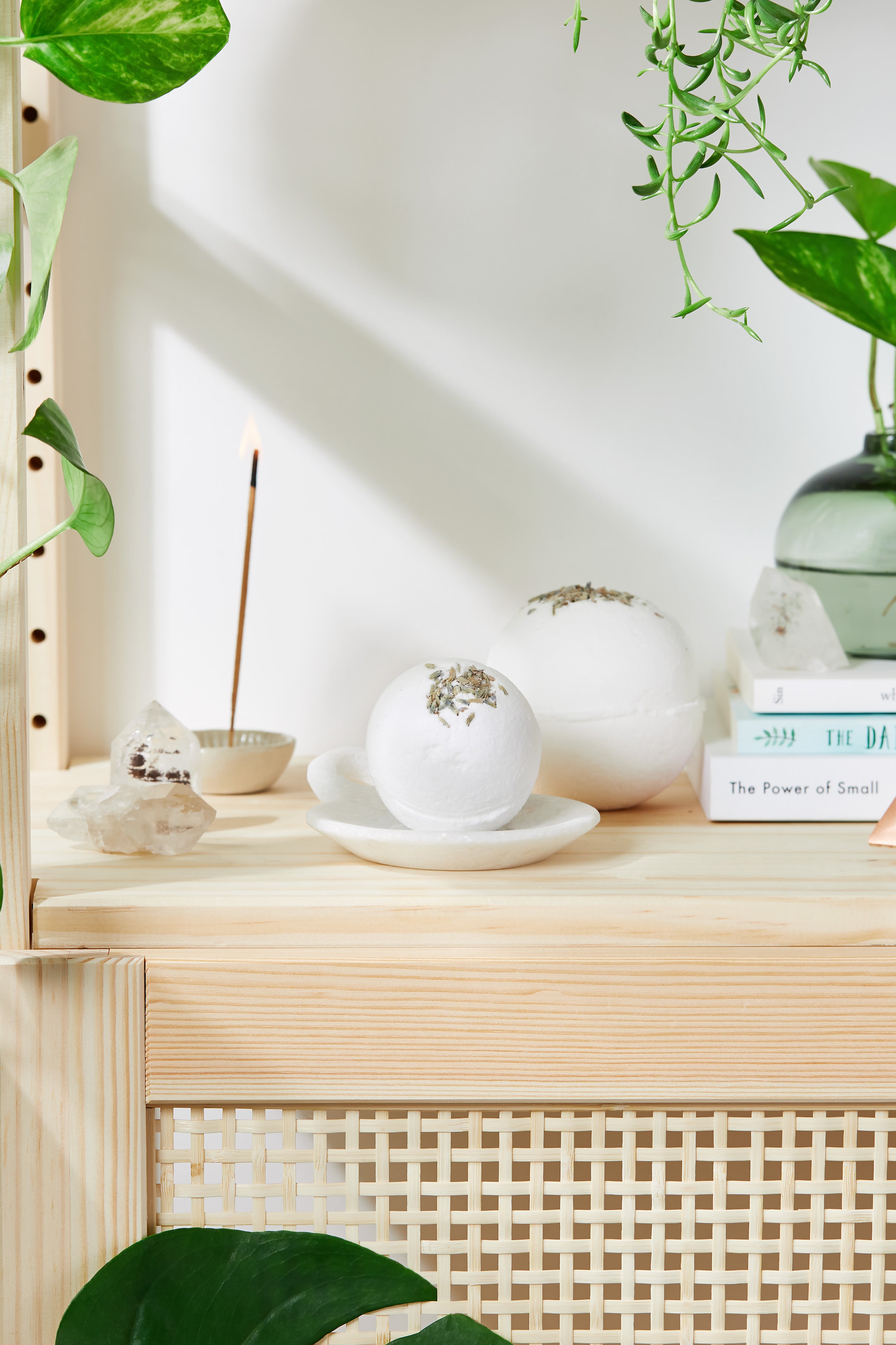 Natural setting with plants, incense, crystals, books, and white bath bombs on a light wooden shelf.