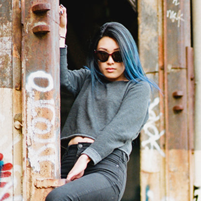 Person with blue hair, sunglasses, gray sweater, and black pants leans on a graffiti-covered wall.