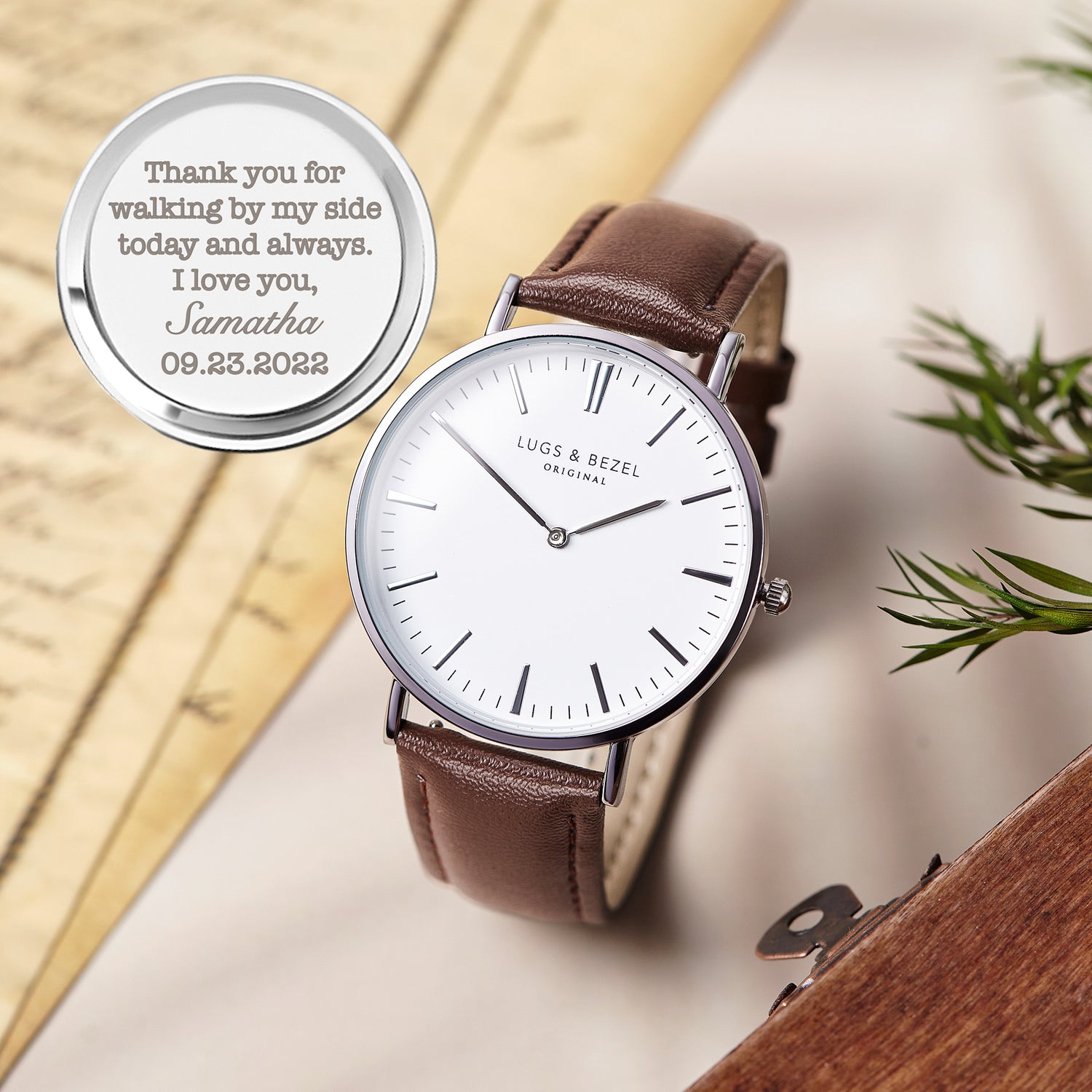 Minimalist watch with brown leather strap, white dial, black markers, and engraved message on the back.