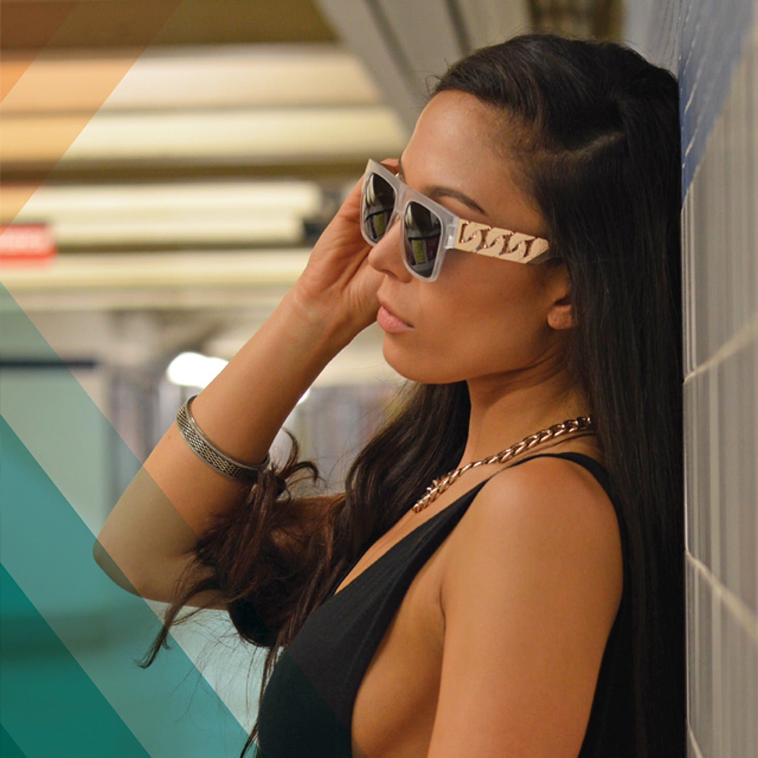 Woman in stylish sunglasses leans against tiled wall, wearing a black top and chain necklace.