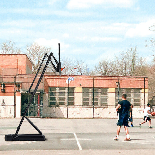 Outdoor basketball court with a bent hoop, red brick building, and people playing under a blue sky.