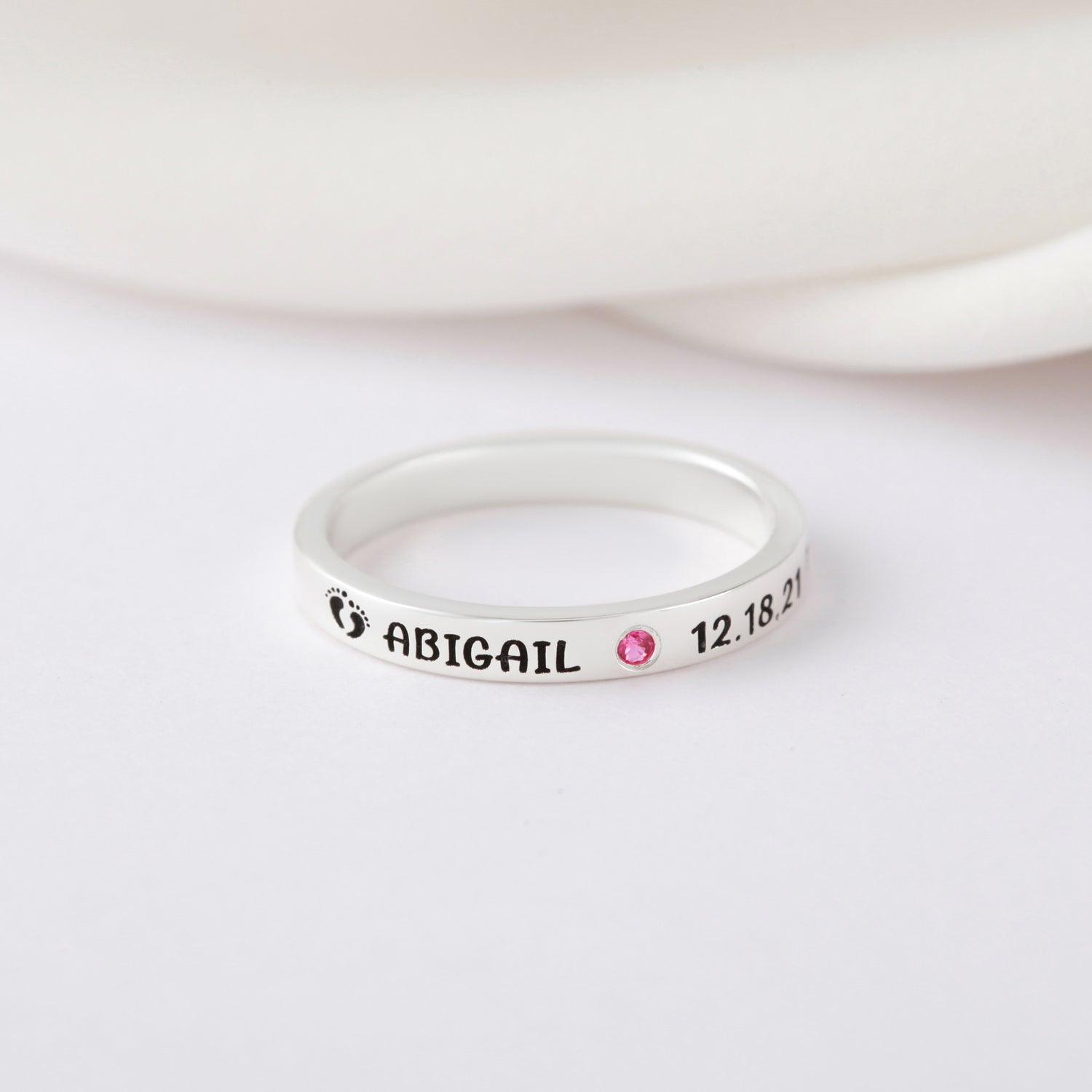 Silver ring with engravings, pink gem, and baby footprints on white background.