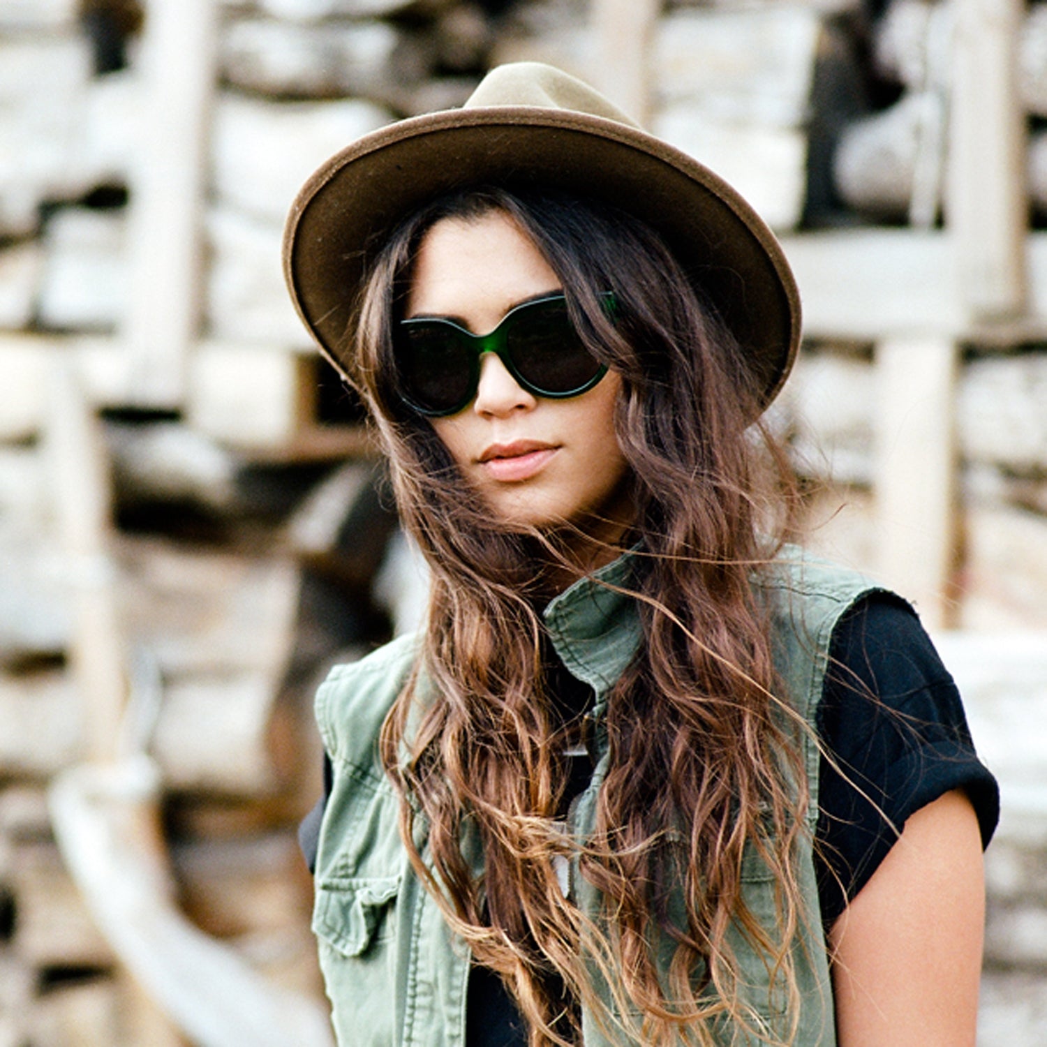 Woman wearing sunglasses, green vest, and hat. Background features stacked wood.