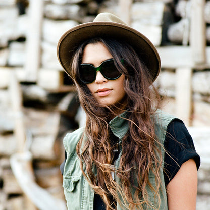 Woman wearing sunglasses, green vest, and hat. Background features stacked wood.