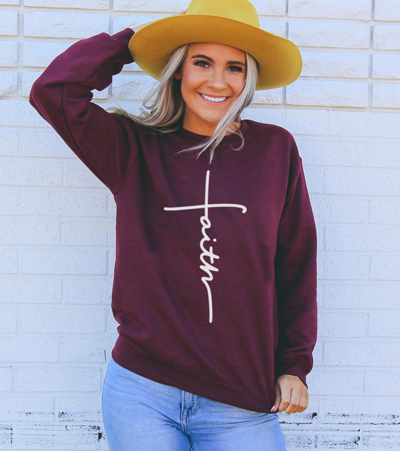 Woman in a burgundy sweater with "faith" text, yellow hat, light jeans against a white brick wall.