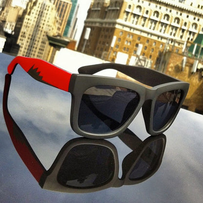 Sunglasses with red arms and black lenses reflecting against a shiny surface, cityscape background.