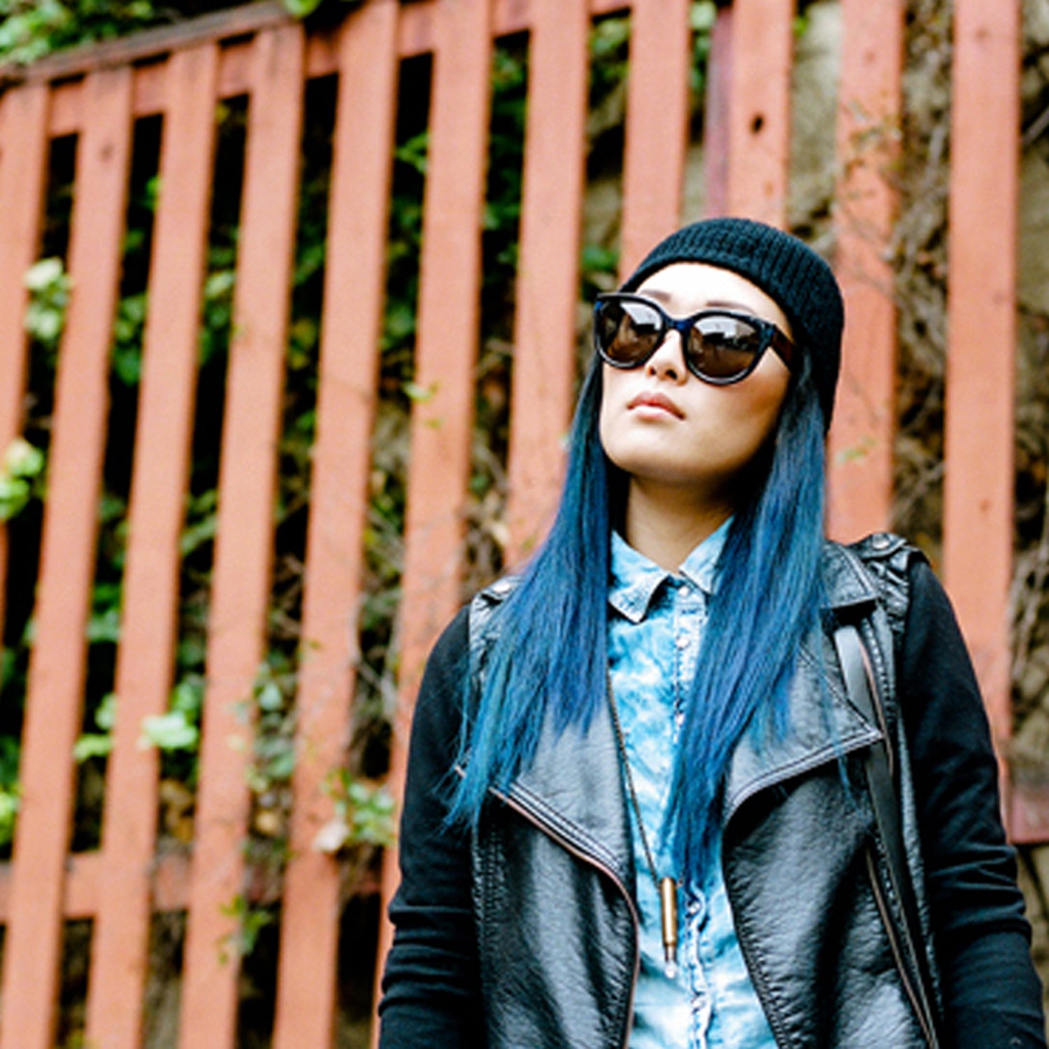Person with blue hair, sunglasses, and a beanie, in front of a wooden fence with greenery.