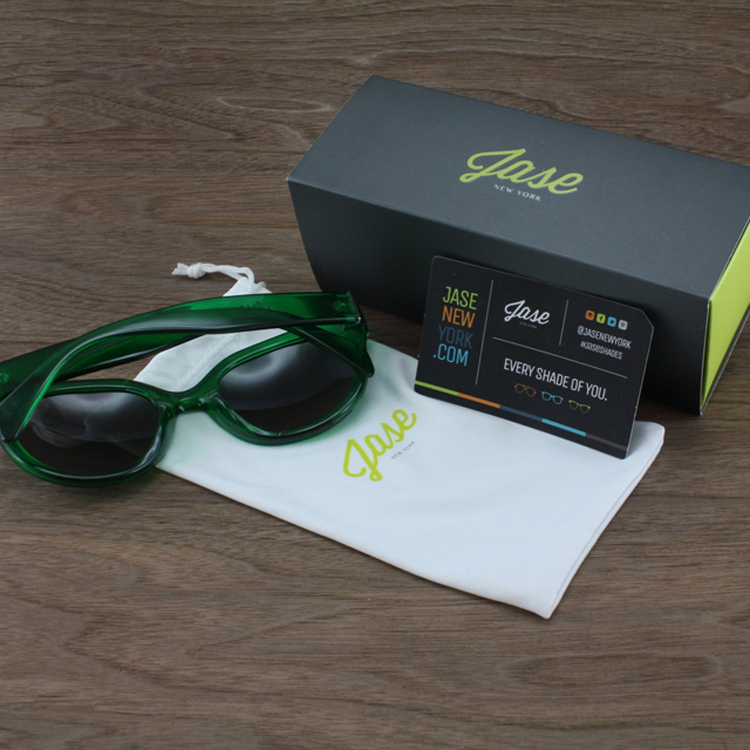 Green sunglasses, a white pouch, black box with "Jase" logo, and a business card on wood surface.