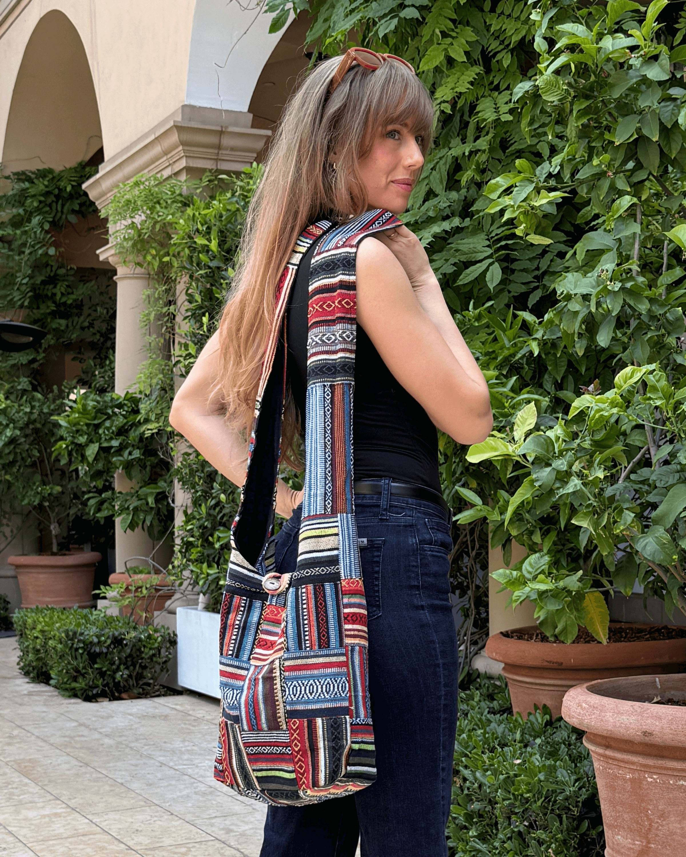 Woman with a colorful patterned tote bag stands in a leafy courtyard, wearing sunglasses on head.