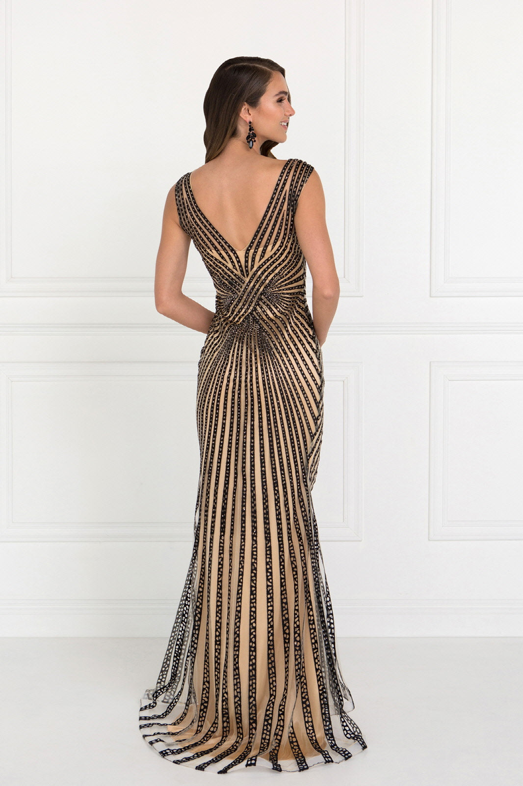 Woman in a black beaded gown with a V-back and intricate vertical pattern stands against a white wall.