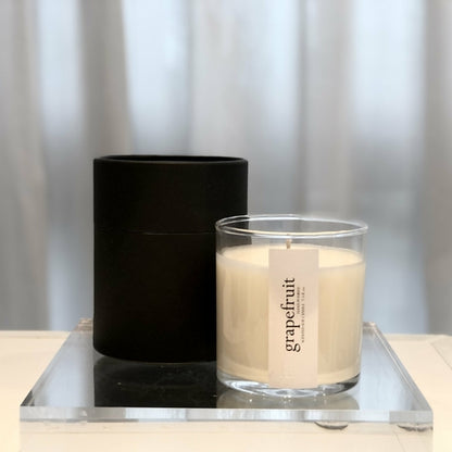 Clear candle labeled "grapefruit" beside black container on reflective surface.