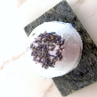 A round bath bomb with lavender buds on top, placed on a gray stone surface.