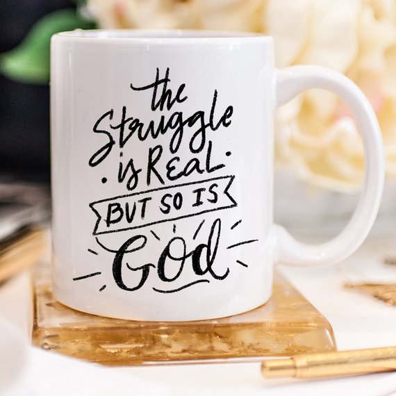 White mug with black cursive text: "The struggle is real, but so is God," on a gold coaster.