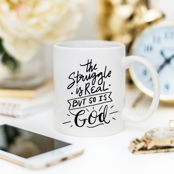 White mug with black text, "The struggle is real, but so is God," near clock and smartphone on desk.