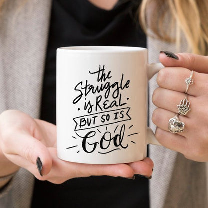 Woman holding white mug with black text: "The struggle is real, but so is God."
