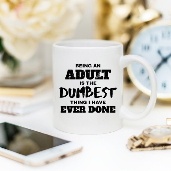 White mug with black text, "Being an adult is the dumbest thing I have ever done," on a desk.