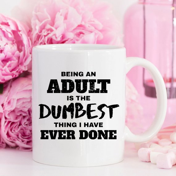 White mug with black text reads "Being an adult is the dumbest thing I have ever done." Pink flowers behind.