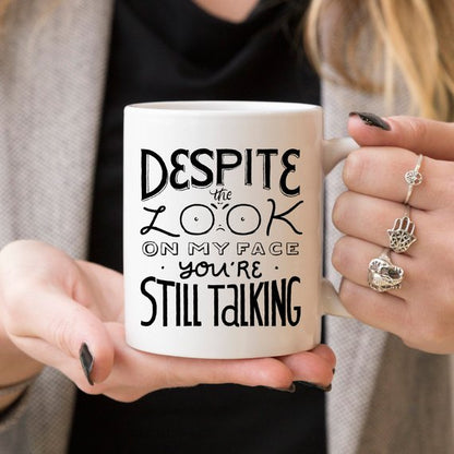 Woman holding a white mug with black text: "Despite the look on my face, you&