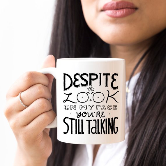 Woman holding a white mug with black text: "Despite the look on my face, you&