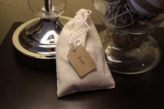 Small white pouch with "Soak" tag on dark table near shiny lamp and glass jar of decor.