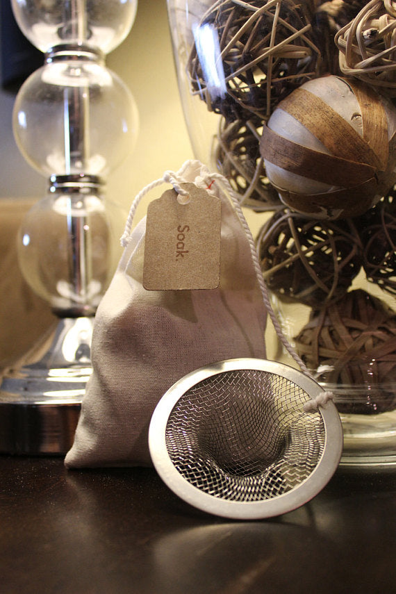 Cloth bag with "Soak" tag, metal mesh strainer, and decorative wicker balls in a glass vase.