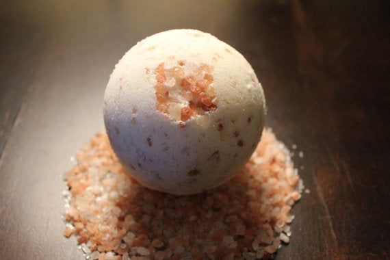 A round, white bath bomb with pink salt crystals sits on a dark surface surrounded by loose salt.