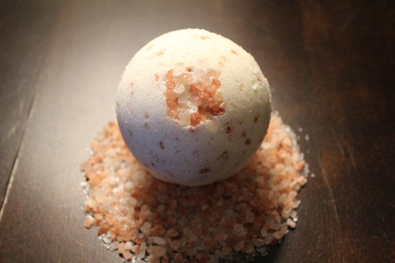 A round bath bomb with pink salt on a wooden surface, surrounded by scattered salt crystals.