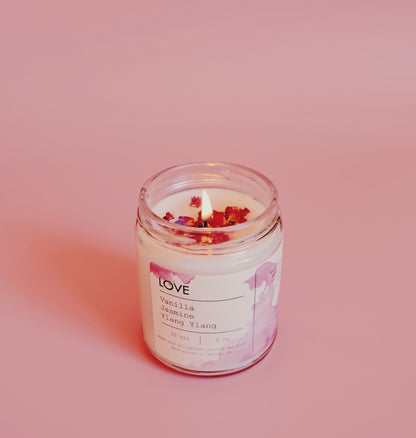 Lit candle in glass jar with label on pink background, topped with red flower petals.