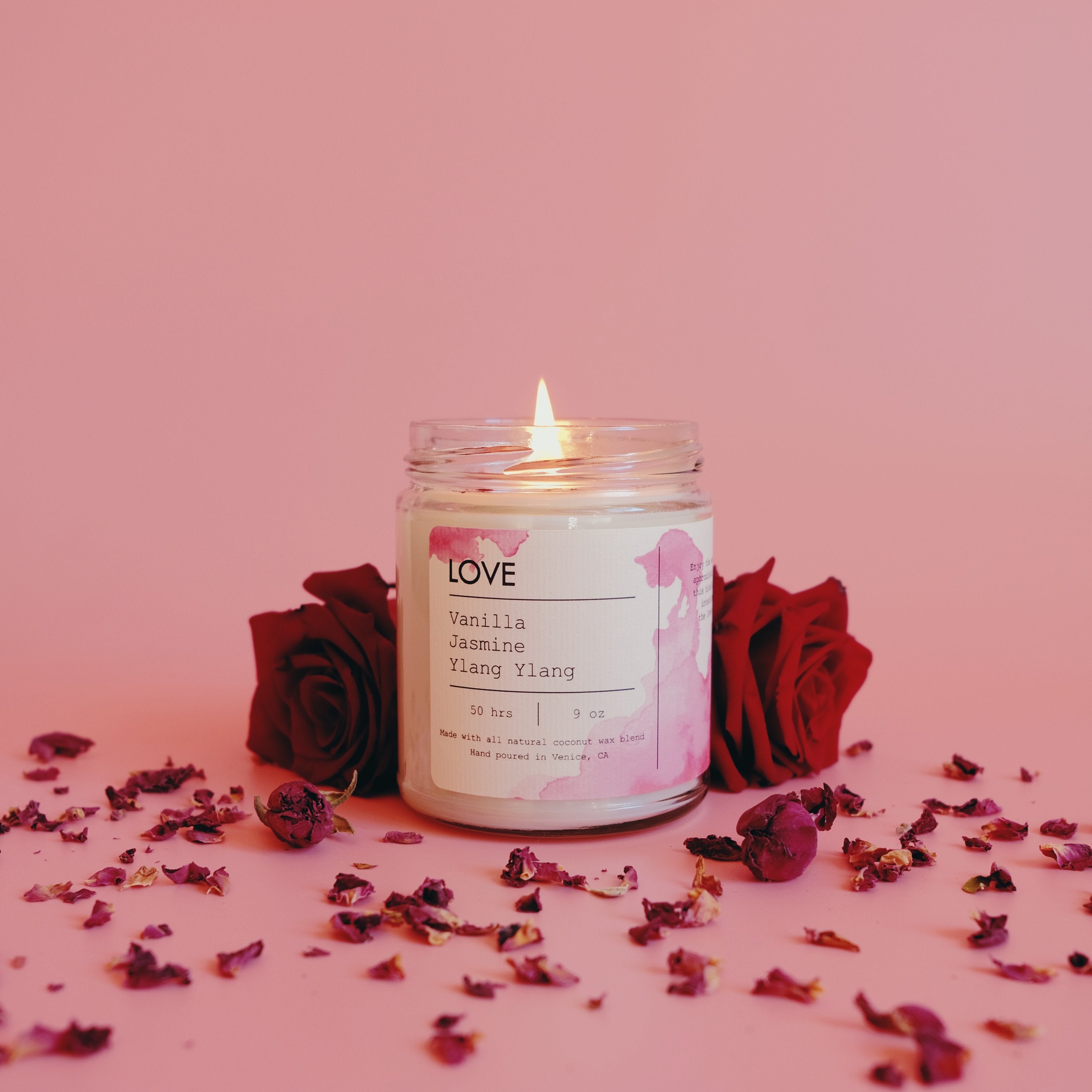 Lit candle in a glass jar with red roses and petals on a pink background.